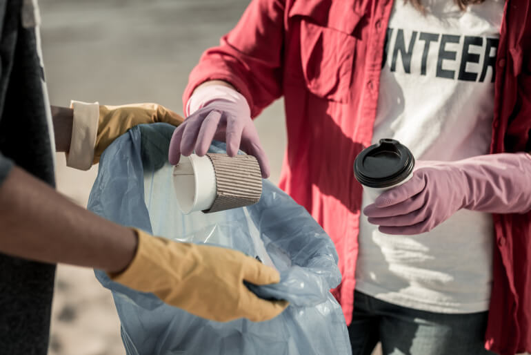 Two people wearing rubber gloves on a beach. One is holding a blue rubbish bag while the other puts empty two coffee cups into the bag, wearing a Volunteer t-shirt.