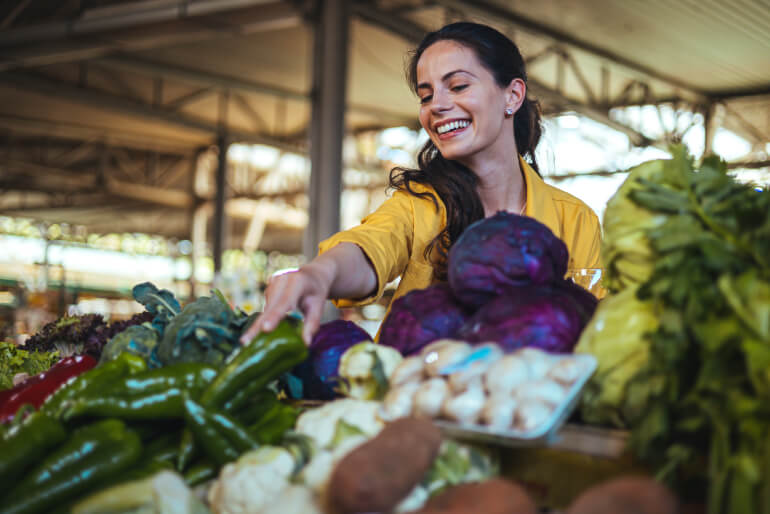 Smiling white woman with dark hair wearing a yellow shirt reaching over a table full of colorful vegetables to grab a green pepper.